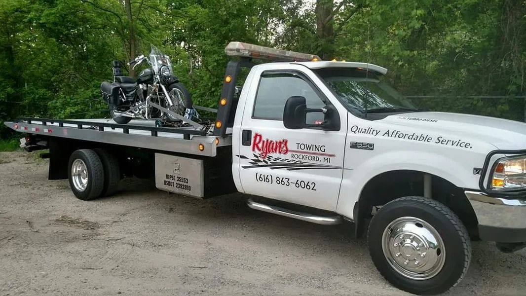 Tow truck with a flatbed carrying a motorcycle, parked on a dirt road near trees.