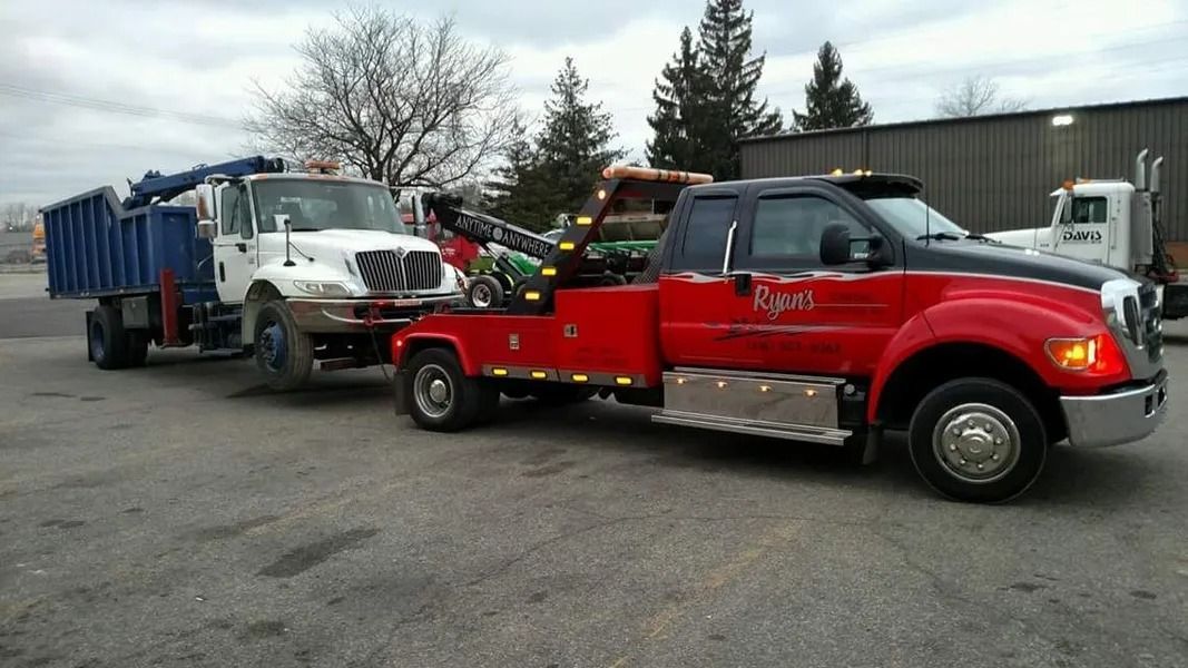 Red tow truck hauling a white truck beside a blue dump truck in an outdoor lot.