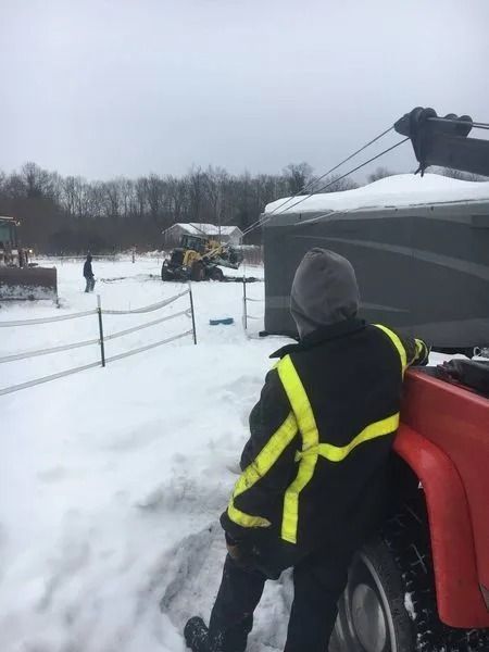 Person in a reflective jacket stands by a red snow vehicle in a snowy field near a cable line.
