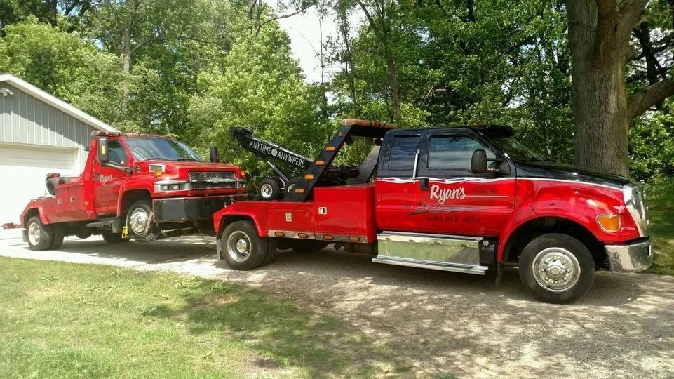 Two red tow trucks parked beside a garage on a grassy driveway, one carrying a vehicle