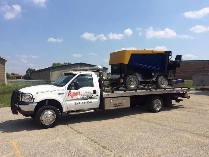 White tow truck carrying a blue-and-yellow truck on a flatbed in a parking lot