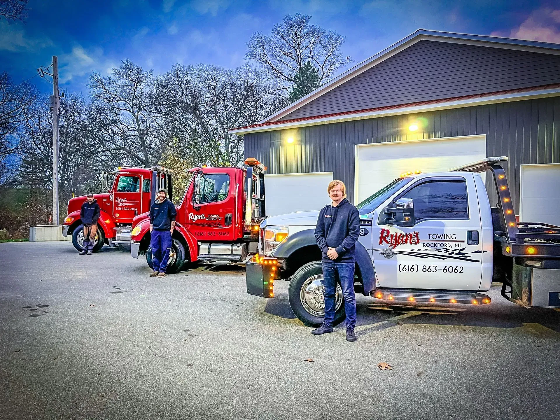Three utility trucks parked by a building at dusk, with two people standing in front.