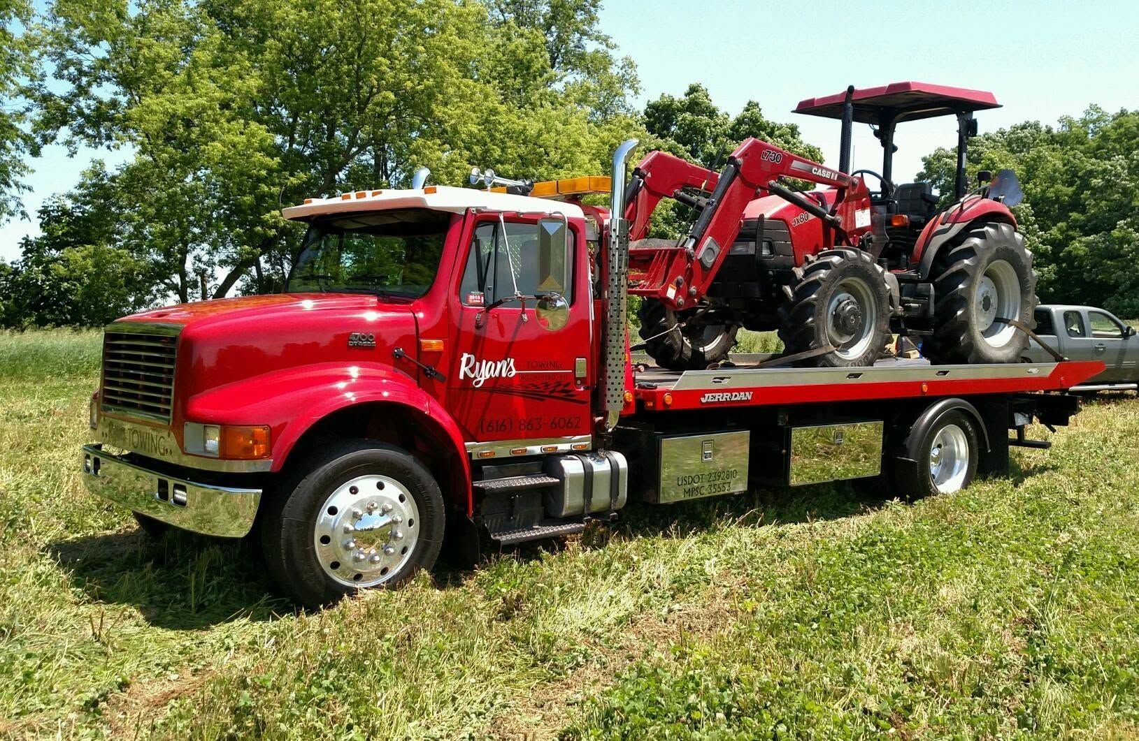 Red flatbed tow truck carrying two red tractors on a grassy field under a blue sky