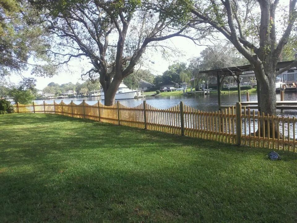 A wooden picket fence lines a green lawn bordering a calm canal with a distant boat and overhanging trees.