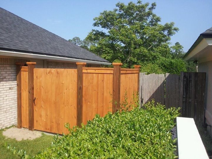 A wooden privacy fence installed between two houses on a sunny day with a green hedge in the foreground.