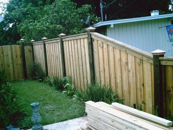 A backyard scene featuring a newly installed wooden privacy fence with decorative post caps and a stack of lumber nearby.