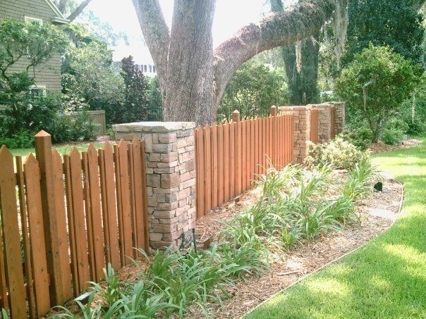 A wooden picket fence with stone pillars runs through a garden with a large tree, shrubs, and a grassy lawn.