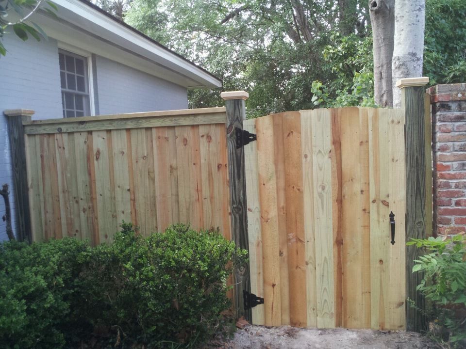 A wooden gate and matching fence panel with black metal hinges installed between two posts near a brick wall and house.
