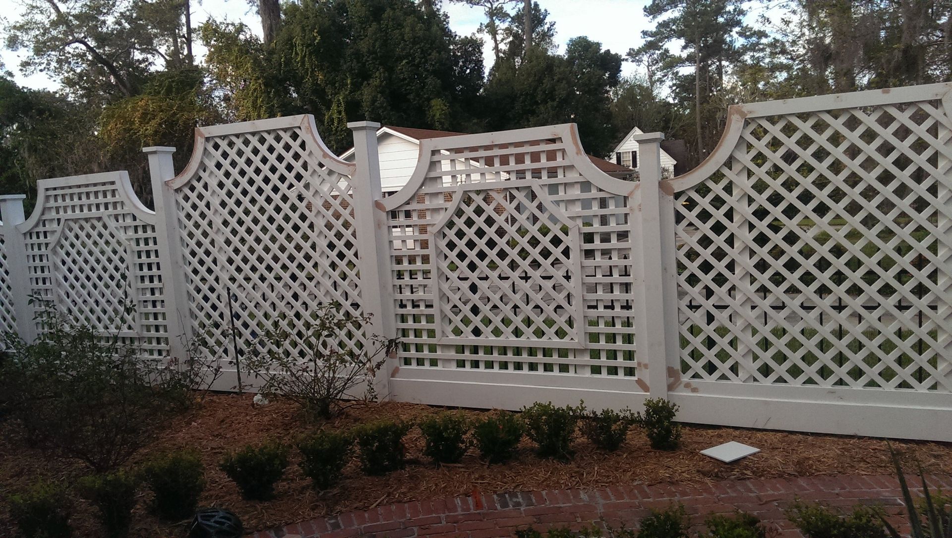 A white, decorative lattice fence stands in a yard with small shrubs planted along the base.