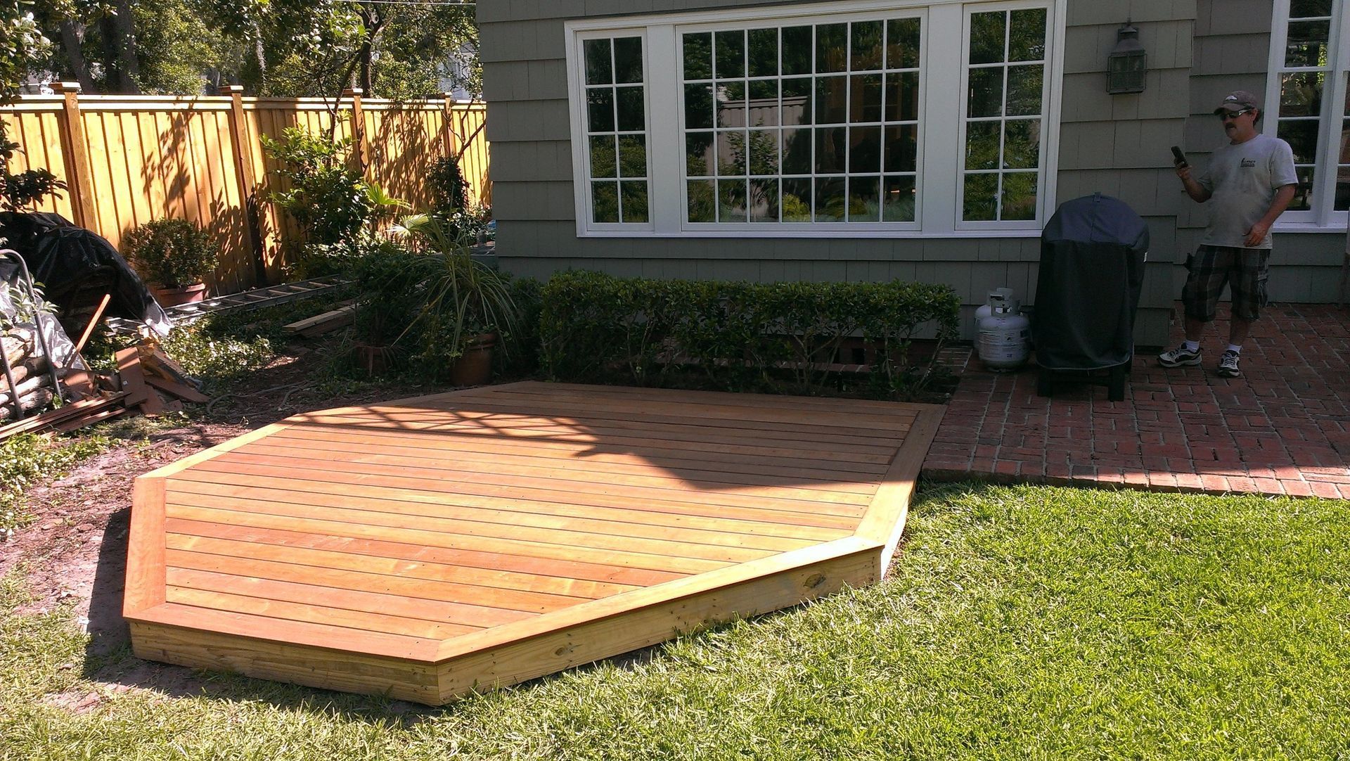 A man stands on a brick patio next to a newly constructed hexagonal wooden deck in a residential backyard.