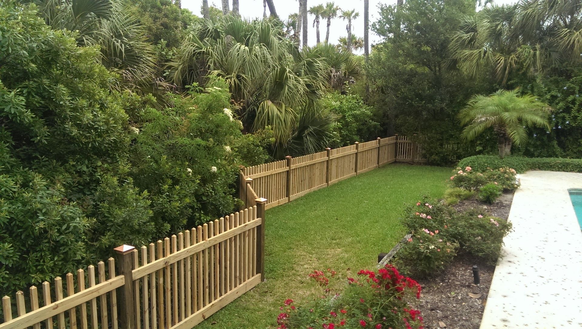 A wooden picket fence lines a green lawn bordering a swimming pool, with lush trees and foliage in the background.