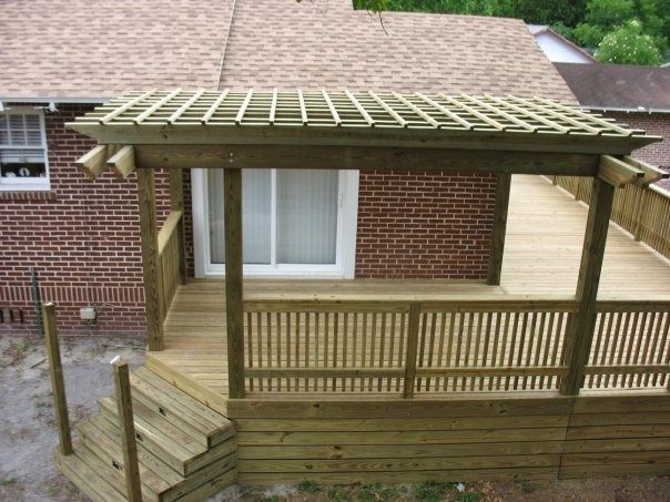 A wooden deck attached to a brick house, featuring a slatted pergola overhead, wooden railings, and stairs leading down.