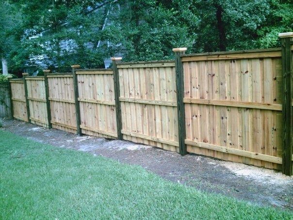 A row of wooden privacy fence panels with dark posts, set against a backdrop of green trees and a grassy lawn.