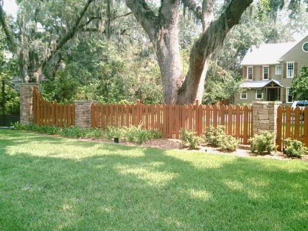 A wooden fence with brick pillars runs across a green lawn in front of a house under large trees.