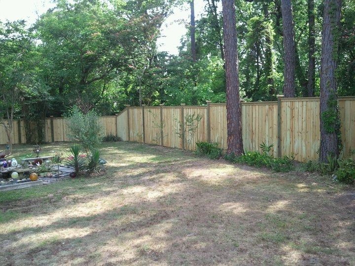 A newly installed wooden privacy fence spans a backyard, with tall trees and greenery visible behind it.