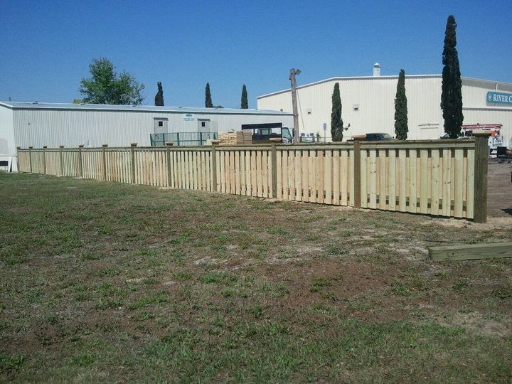 A newly installed wooden picket fence stands on a grassy field in front of a light-colored commercial building.