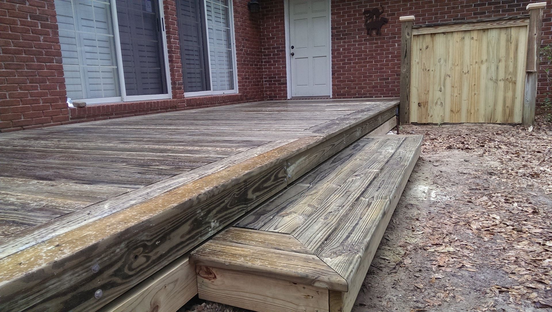 A wooden deck with a set of stairs leading down to a dirt ground area next to a brick house with a white door.