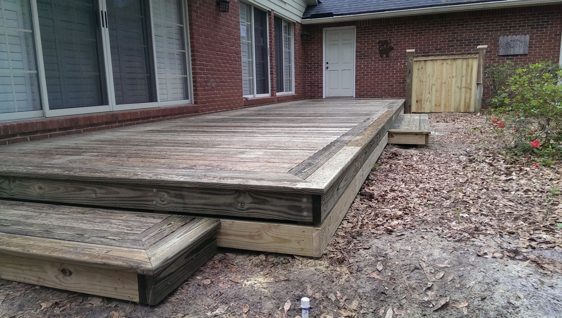 A wooden deck attached to a brick house exterior, featuring a small step and surrounded by dry leaves on the ground.