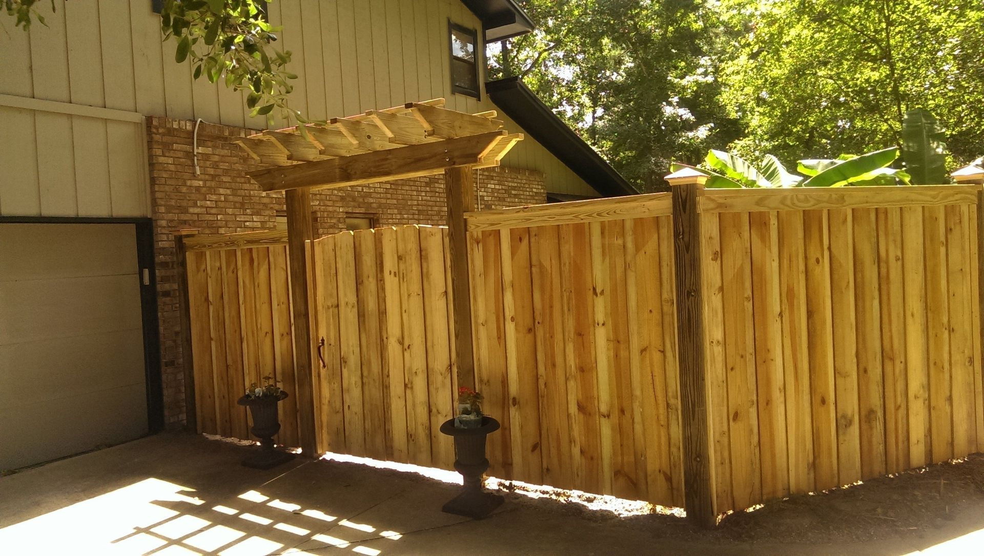 A wooden fence with an integrated pergola-style gate entrance stands in front of a house on a sunny day.