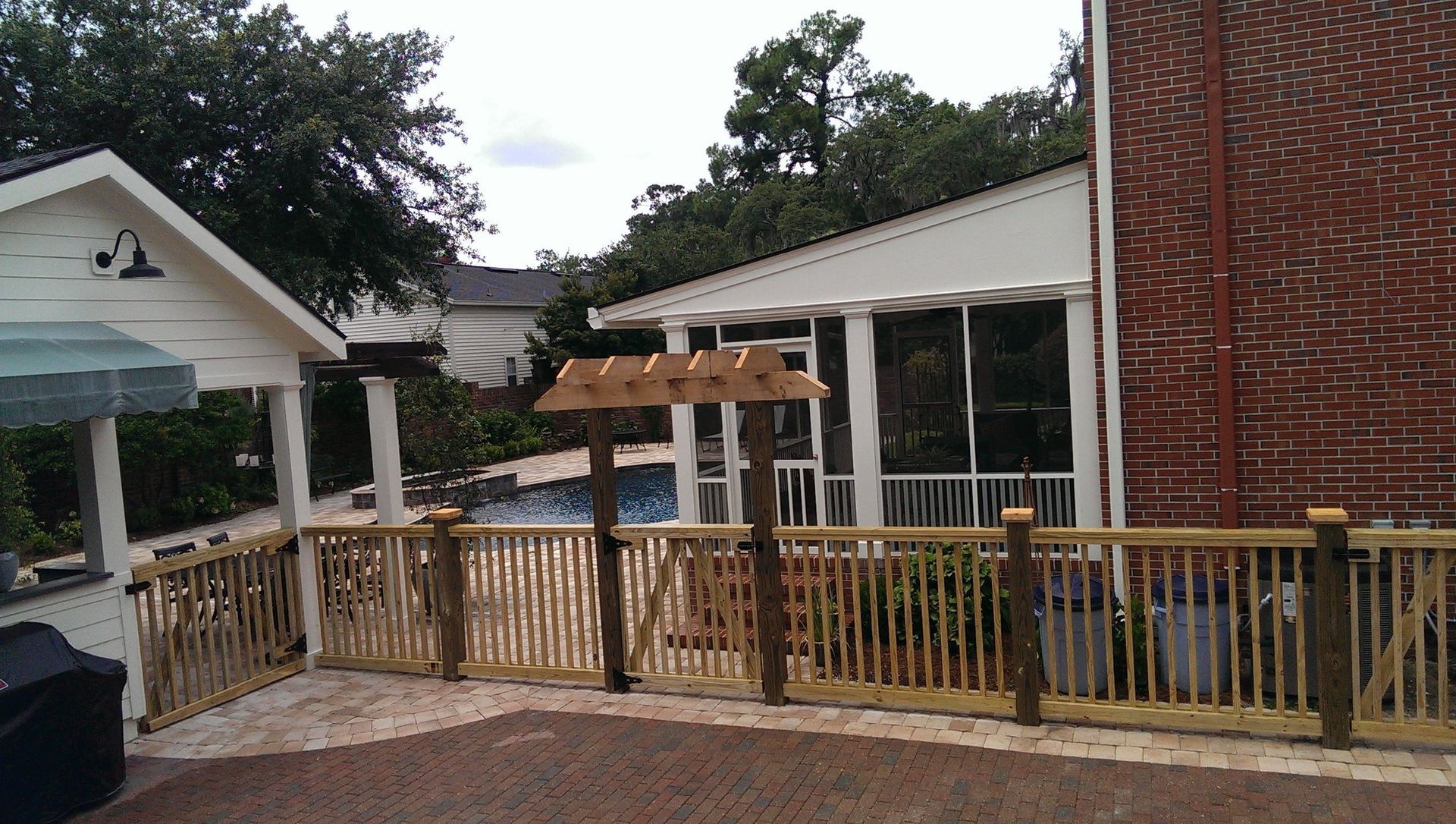 A wooden deck fence with a gate and a small decorative arbor, positioned in front of a house with a pool and patio.
