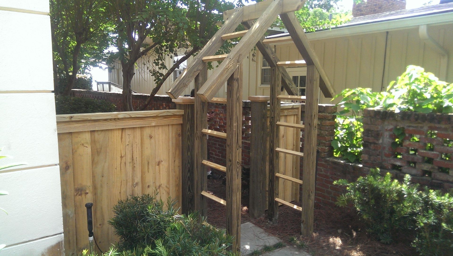 A wooden garden arbor with a ladder-like frame stands between a wooden fence and a brick wall in a sunny backyard.