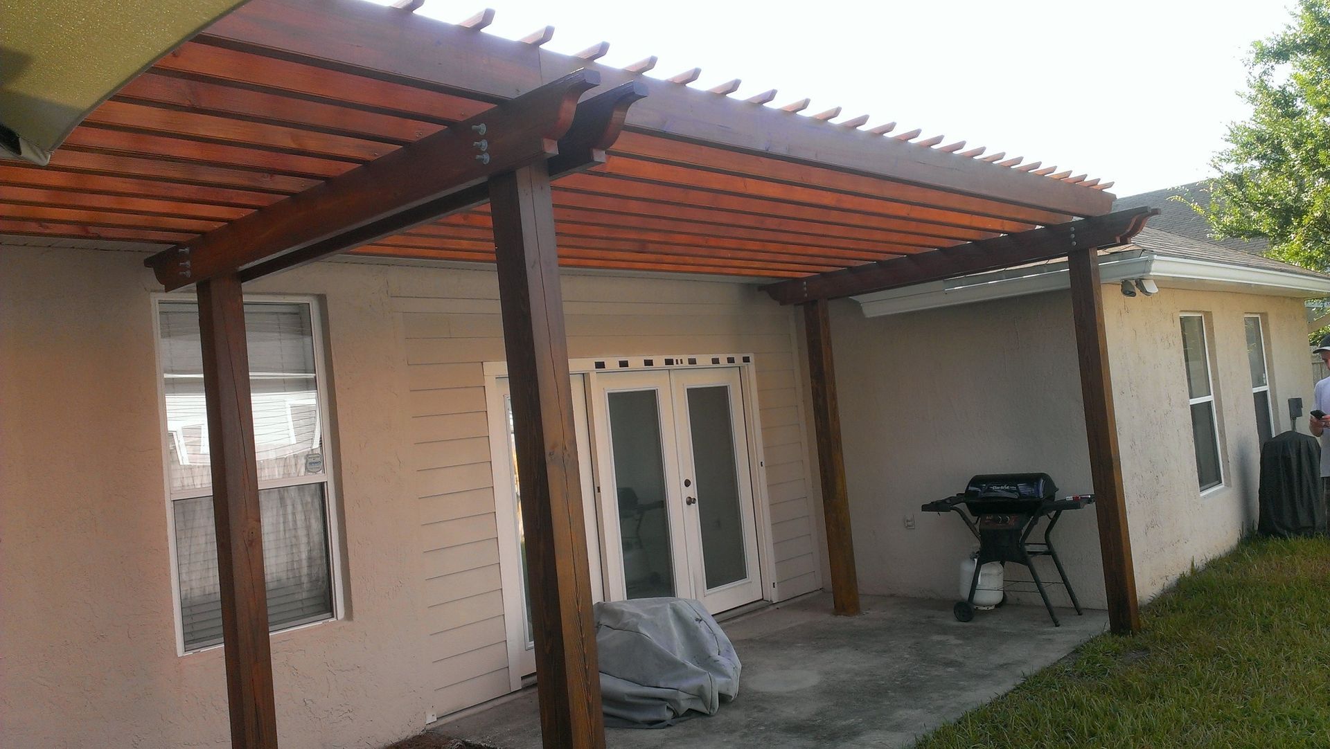 A wooden pergola attached to a beige house, covering a concrete patio with a grill and outdoor furniture.