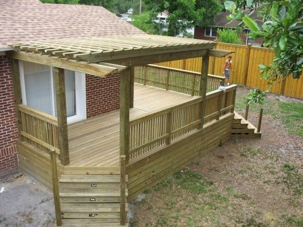 A wooden deck attached to a brick house, featuring a slatted pergola cover and stairs leading to a backyard.