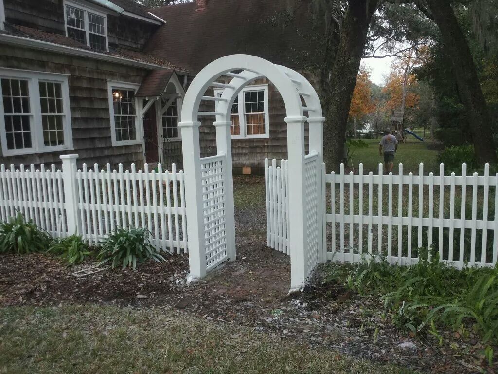 A white picket fence with an arched gate in front of a rustic, shingled house surrounded by trees.