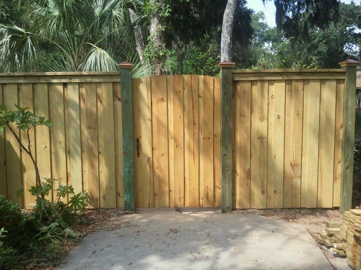 A light wood fence with a matching arched gate positioned between two green wooden posts on a concrete surface.