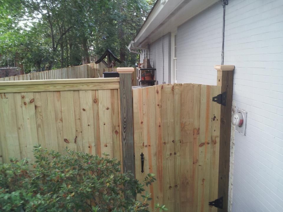 A wooden gate connected to a house exterior wall and a fence, with a green bush in the foreground.