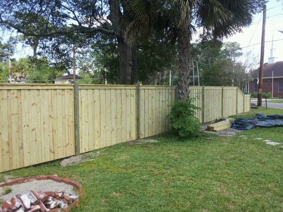 A new, light-colored wooden privacy fence stands in a grassy yard, partially behind a tree and near a brick fire pit.