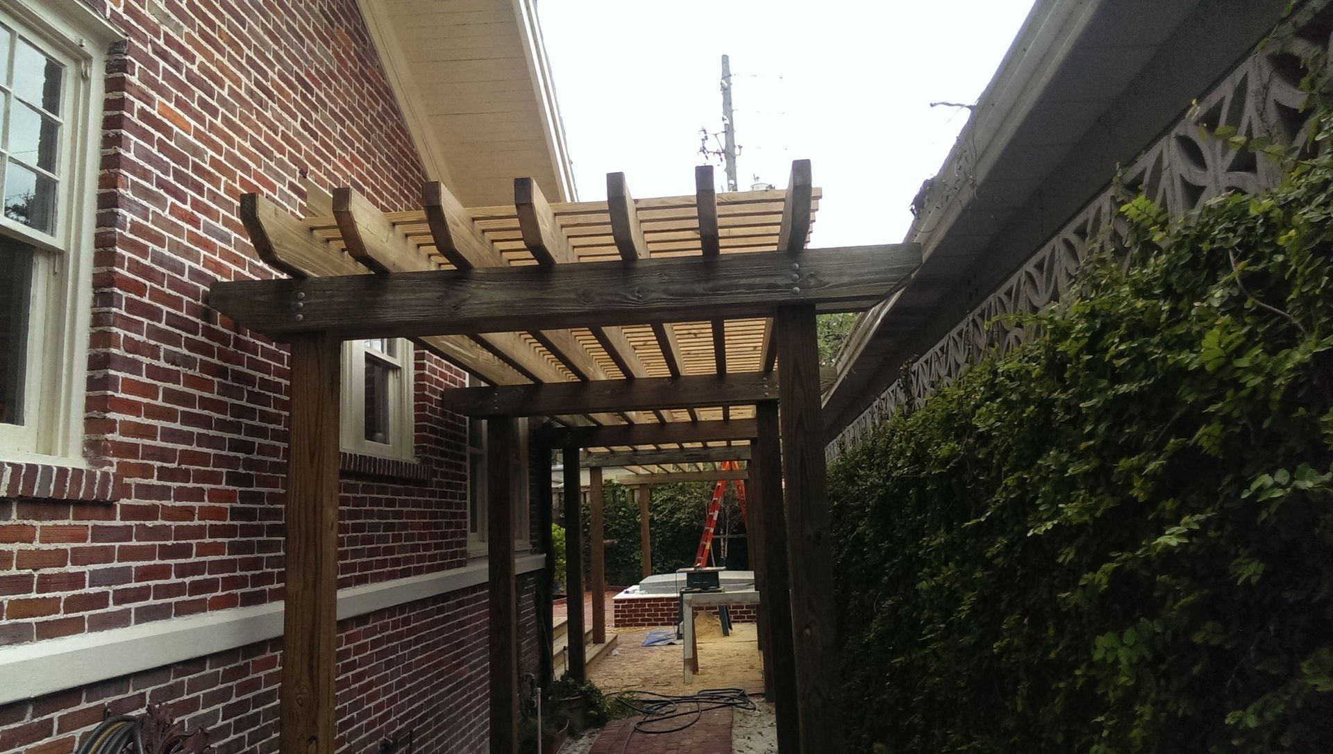 A wooden pergola structure built in a narrow outdoor walkway between a brick house wall and a leafy green hedge.