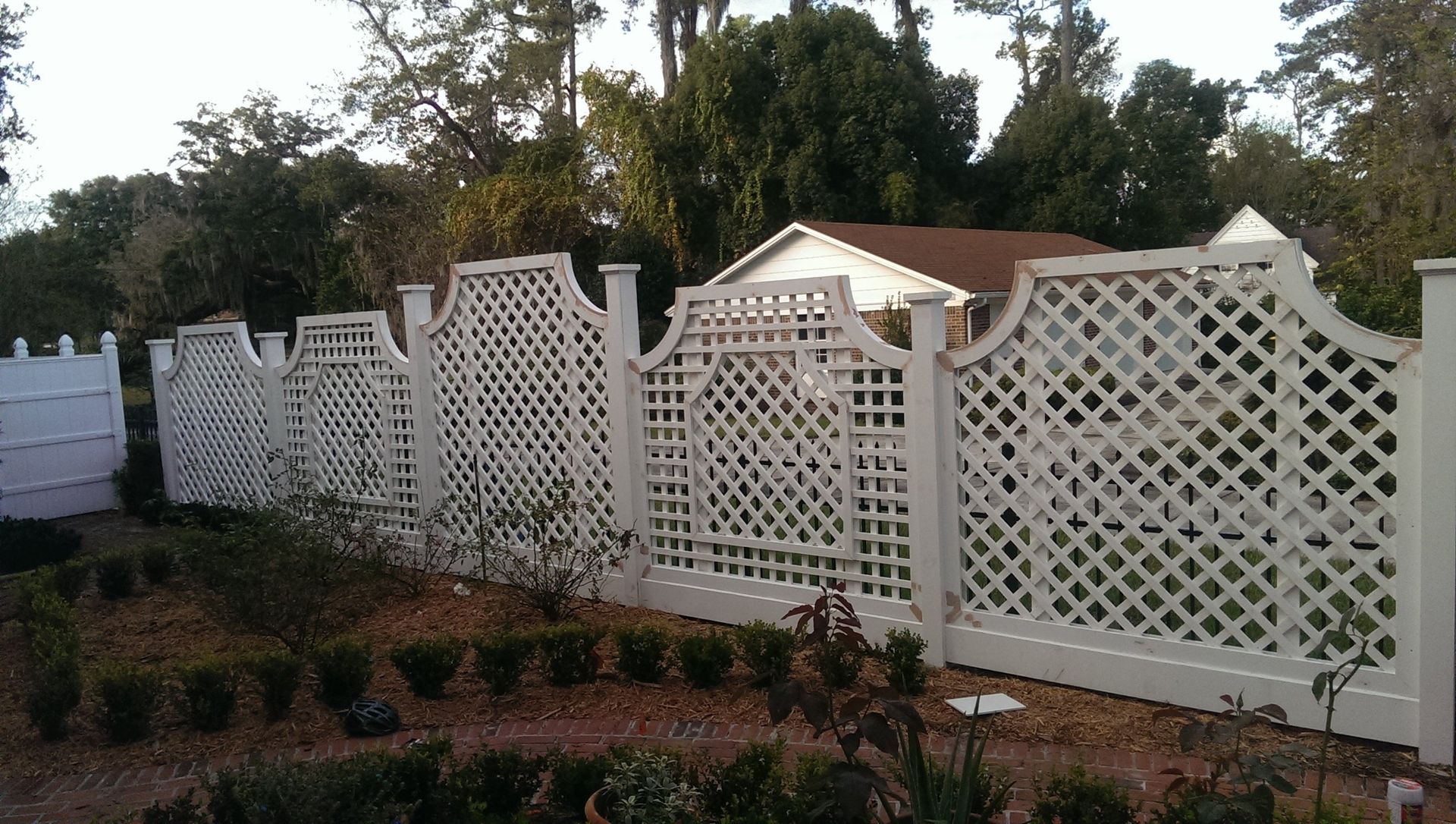 A white decorative lattice fence with arched sections runs along a garden bed of small shrubs, with trees in the background.