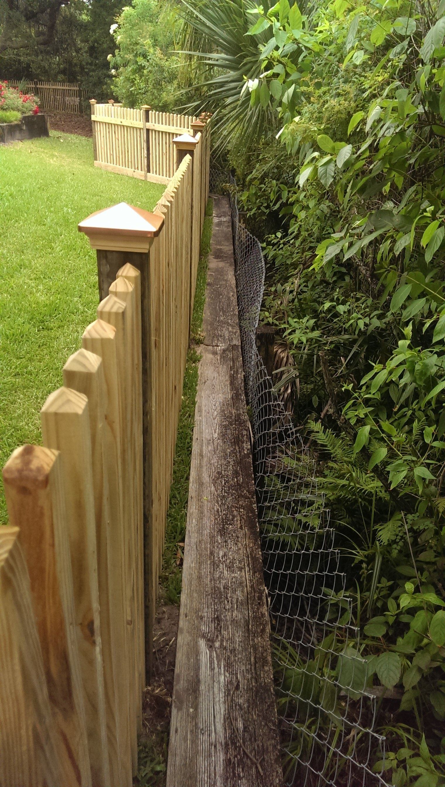 A wooden fence stands next to a weathered timber wall, separating a grassy lawn from a dark, overgrown drainage area.