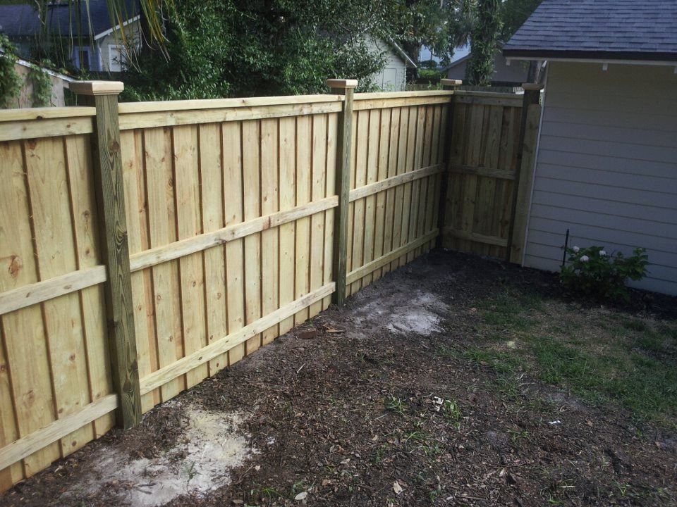 A newly installed light-wood privacy fence borders a backyard next to the side of a light-colored house.