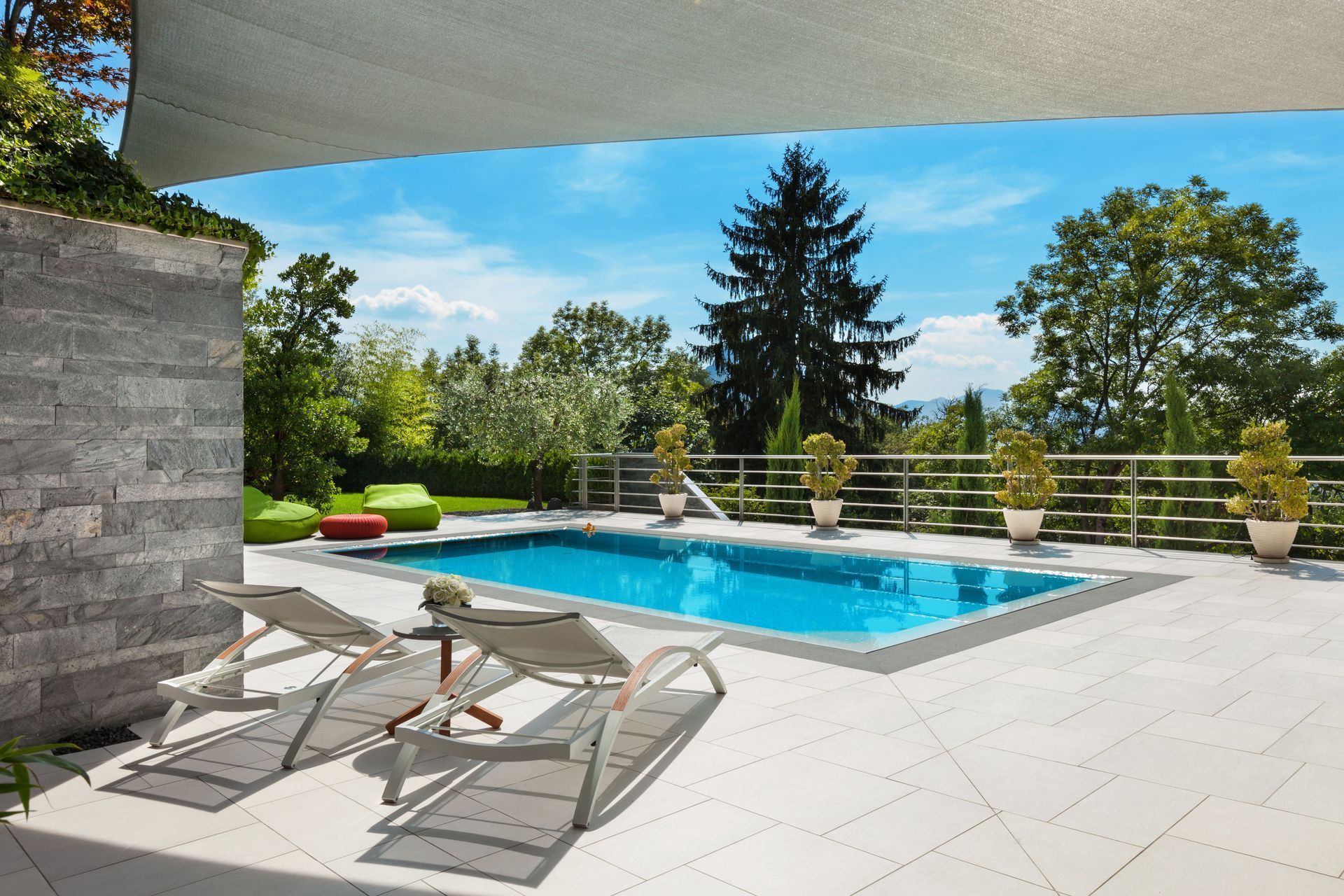 Modern patio with lounge chairs and a swimming pool, surrounded by trees and a glass railing.