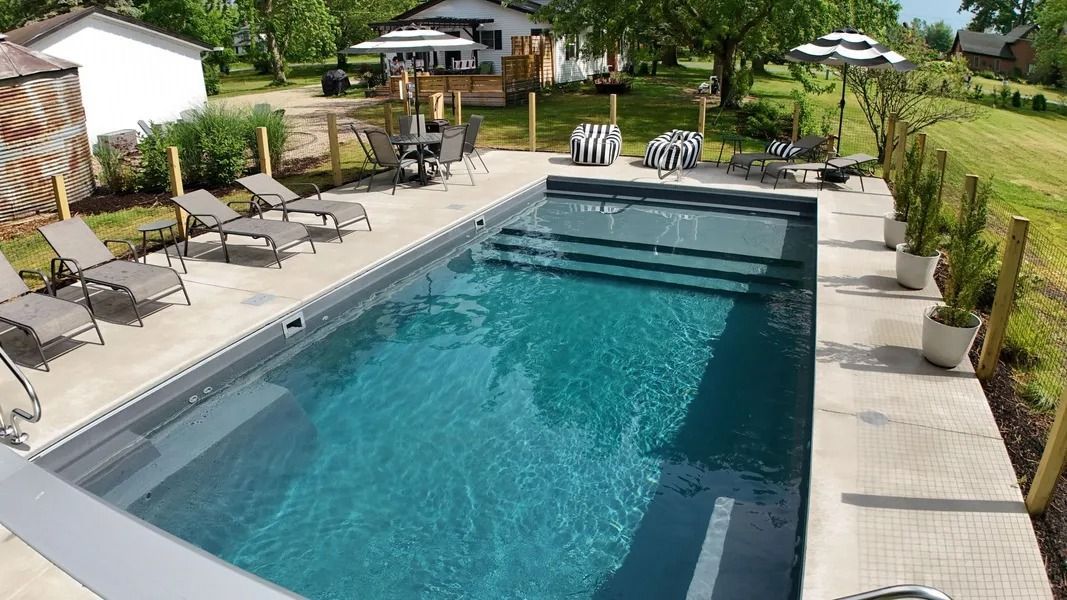 Backyard swimming pool with lounge chairs, umbrellas, and a paved deck on a sunny day