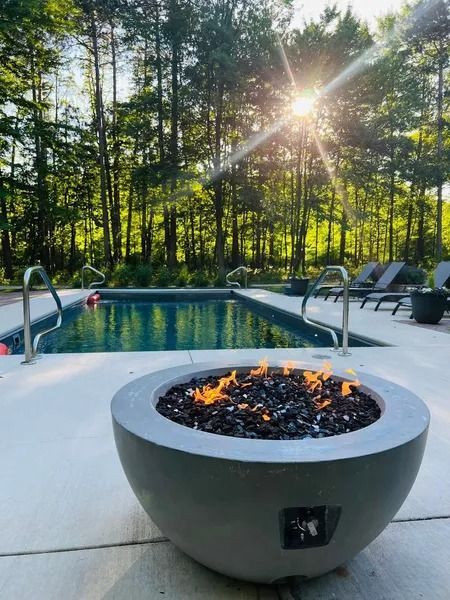 Backyard pool with a lit fire bowl in the foreground, lounge chairs, and sunlit trees behind it