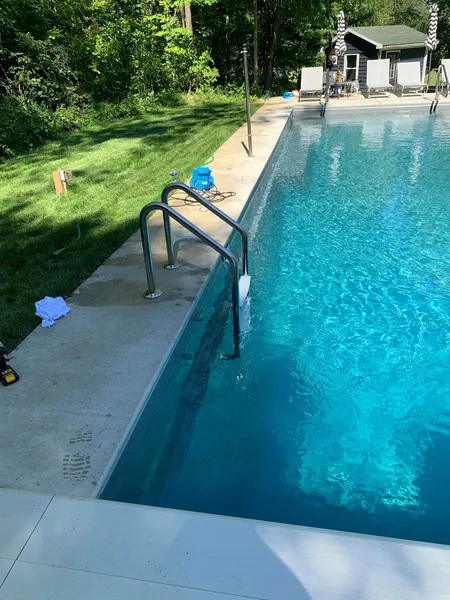 Outdoor swimming pool with a metal handrail, concrete deck, and trees in the background.