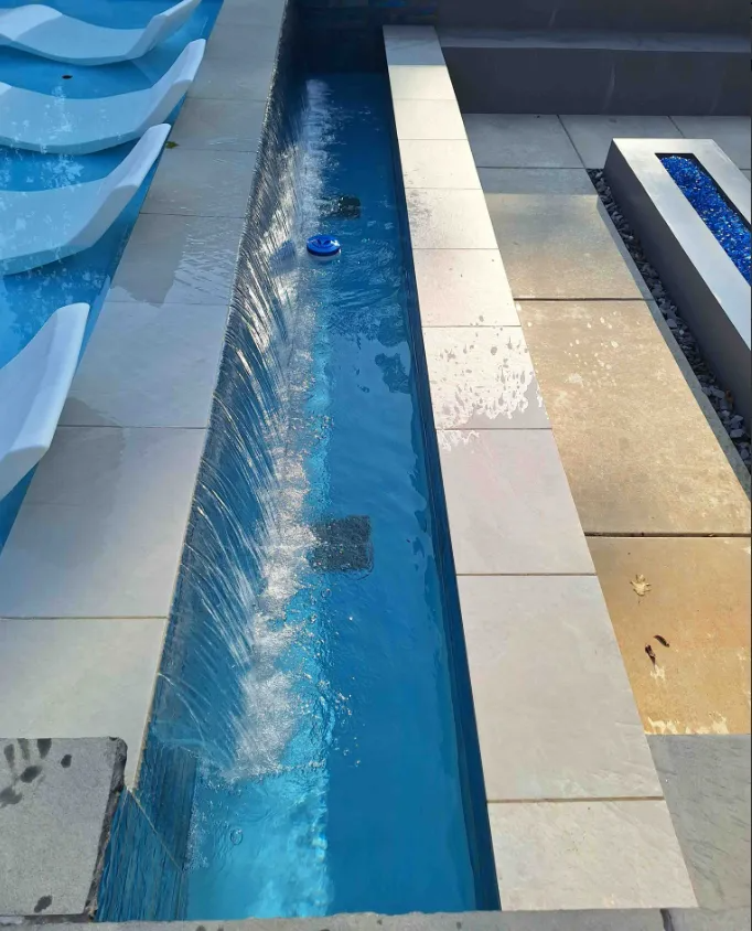 Narrow blue pool channel beside tiled deck and lounge chairs, with water ripples and a small floating object