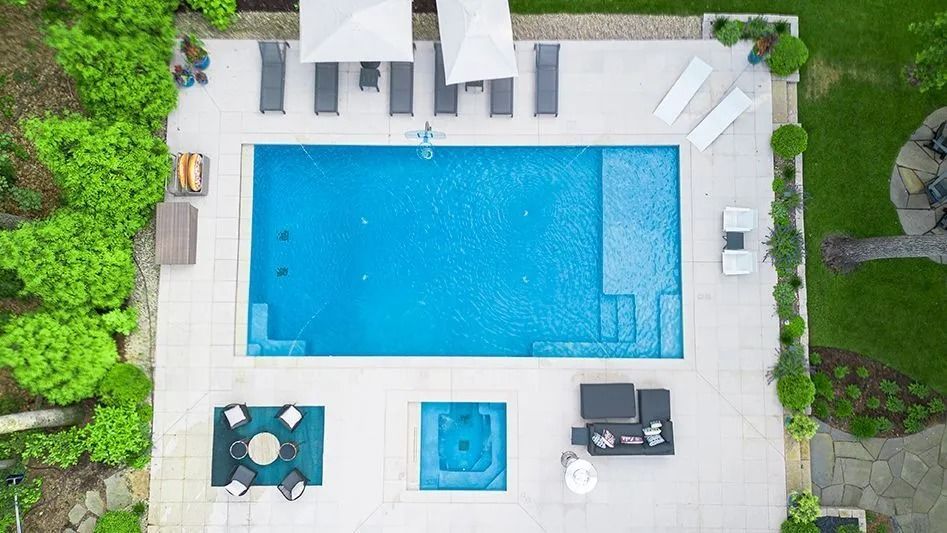 Overhead view of a backyard pool with lounge chairs, umbrellas, and a hot tub on a white patio