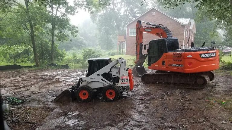 A Bobcat and an excavator in a muddy construction site during a rainstorm; a house is in the background.