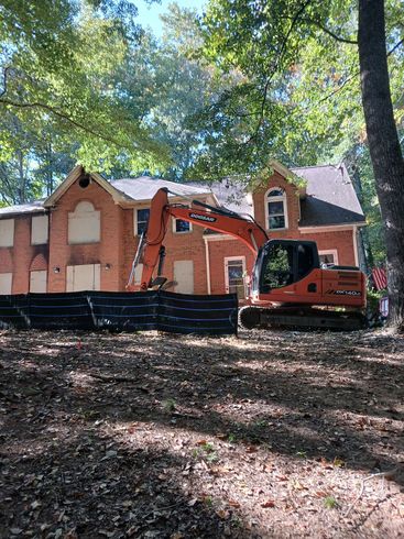 An excavator tearing down a two-story brick building. The windows are boarded up, and leaves cover the ground.