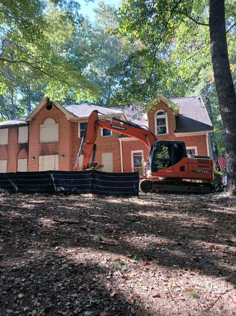 An excavator tearing down a two-story brick building. The windows are boarded up, and leaves cover the ground.