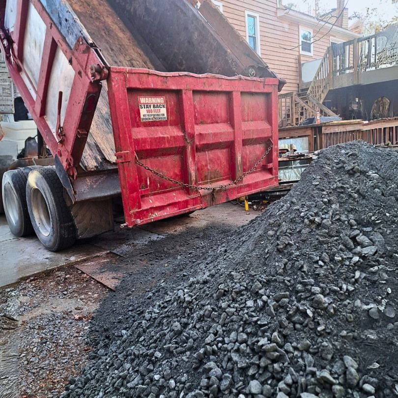 Dump truck unloading gravel onto a driveway near a house and wooden deck. Red truck, gray gravel.