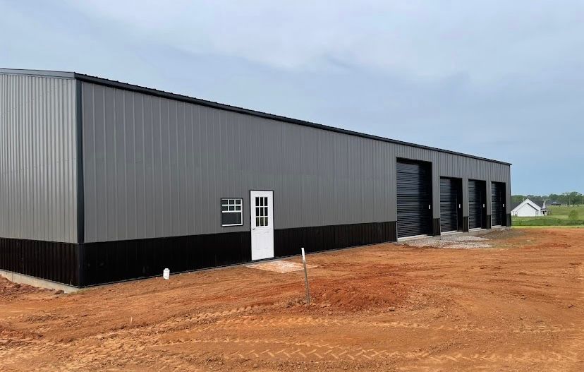 Large gray and black metal industrial building with garage doors and a white door, set in a dirt lot under a cloudy sky.