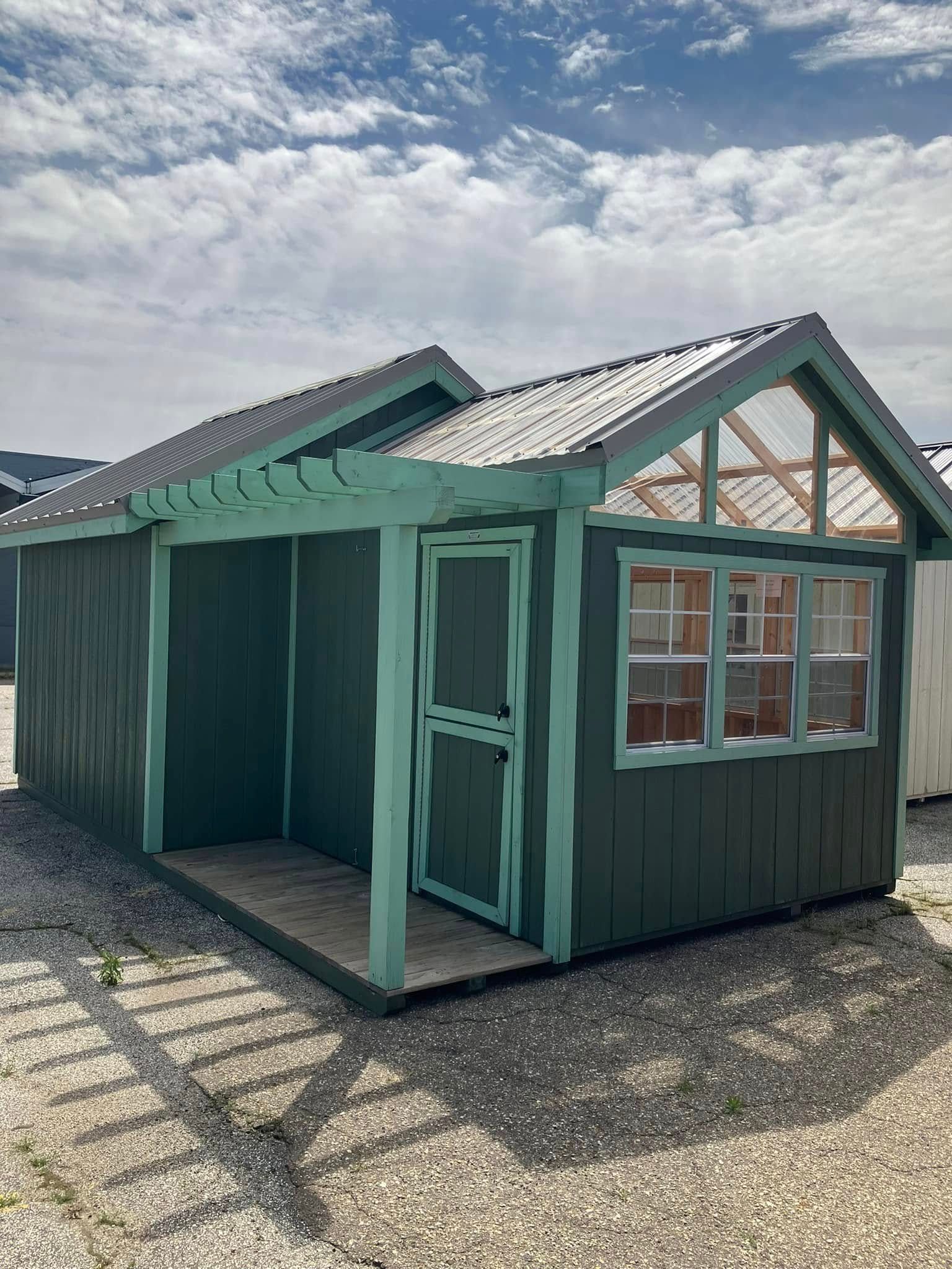 A green shed with a pergola and windows is sitting on top of a gravel lot.