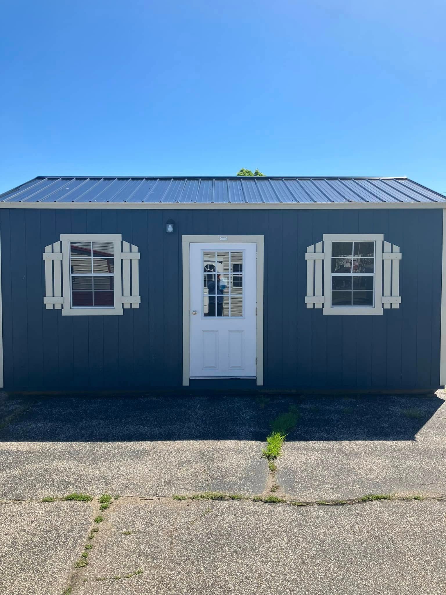 A small shed with two windows and a door is sitting on top of a gravel driveway.