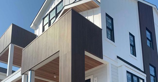 Modern apartment building exterior with white and gray facade and wooden balcony accents against a blue sky.
