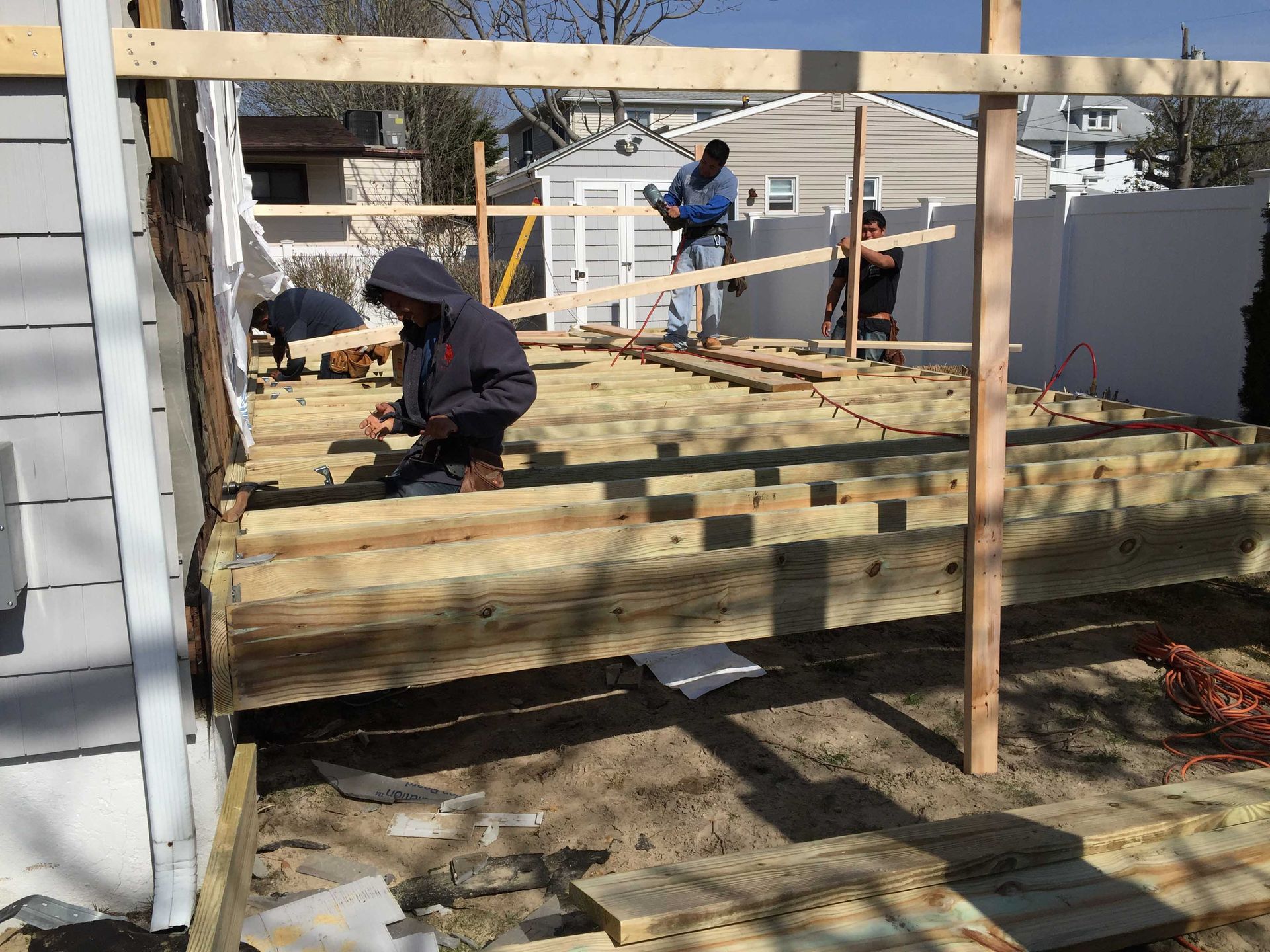 Workers framing a wooden deck in a backyard, with exposed joists and lumber supports under construction